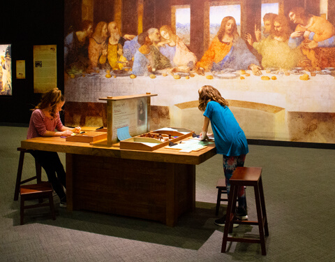 Two girls at a table writing in front of the Last Supper.