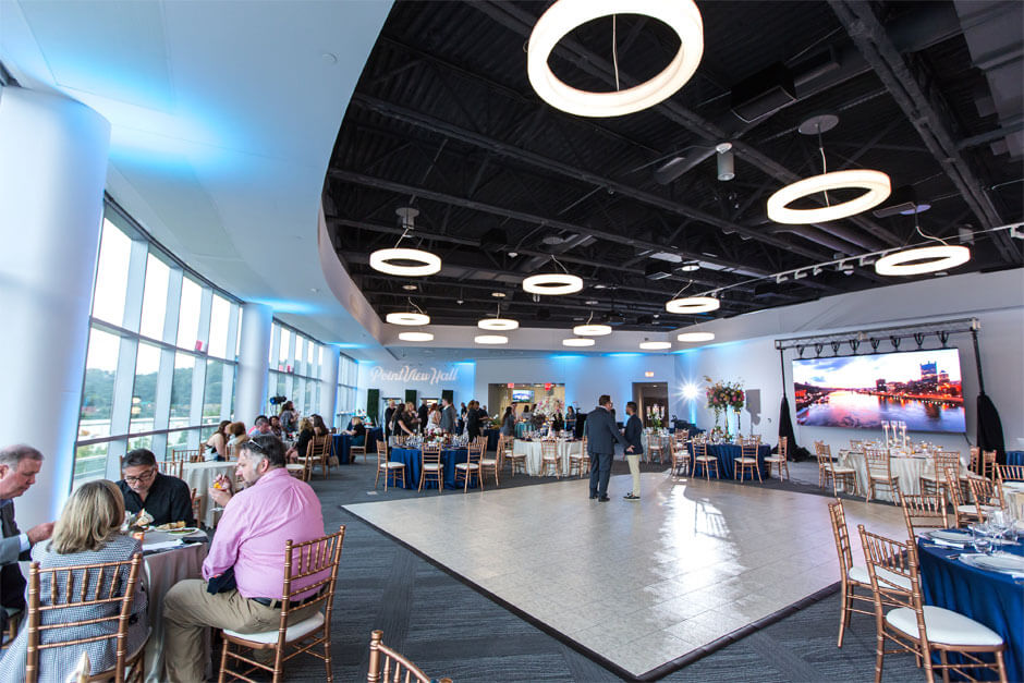 Interior of PointView Hall, tables, chairsm and dance floor. Men and women in business attire are sitting and eating at round tables. There is a presentation screen showing a daytime picture of the Pittsburgh Skyline.